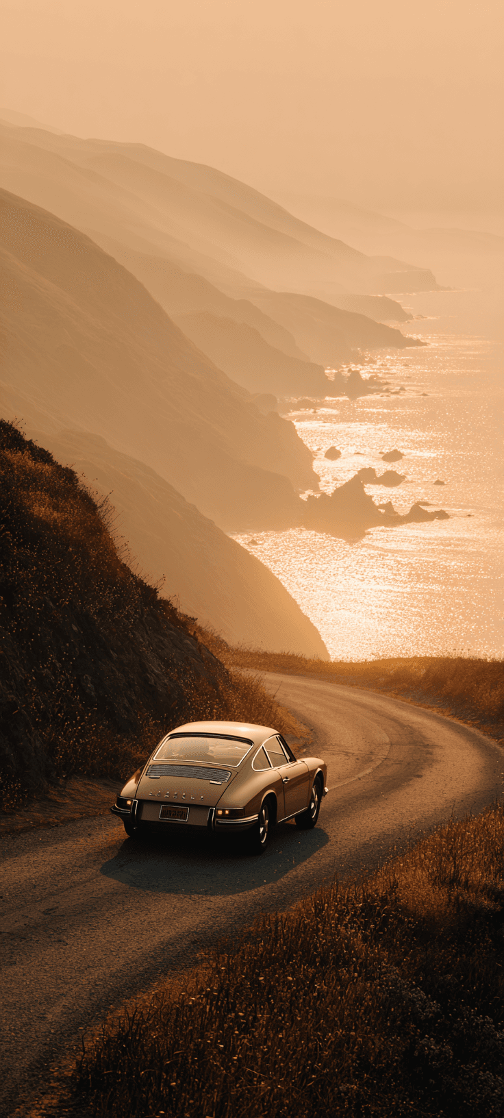 Vintage car driving on a winding coastal road during a golden sunset with ocean and cliffs in the background.