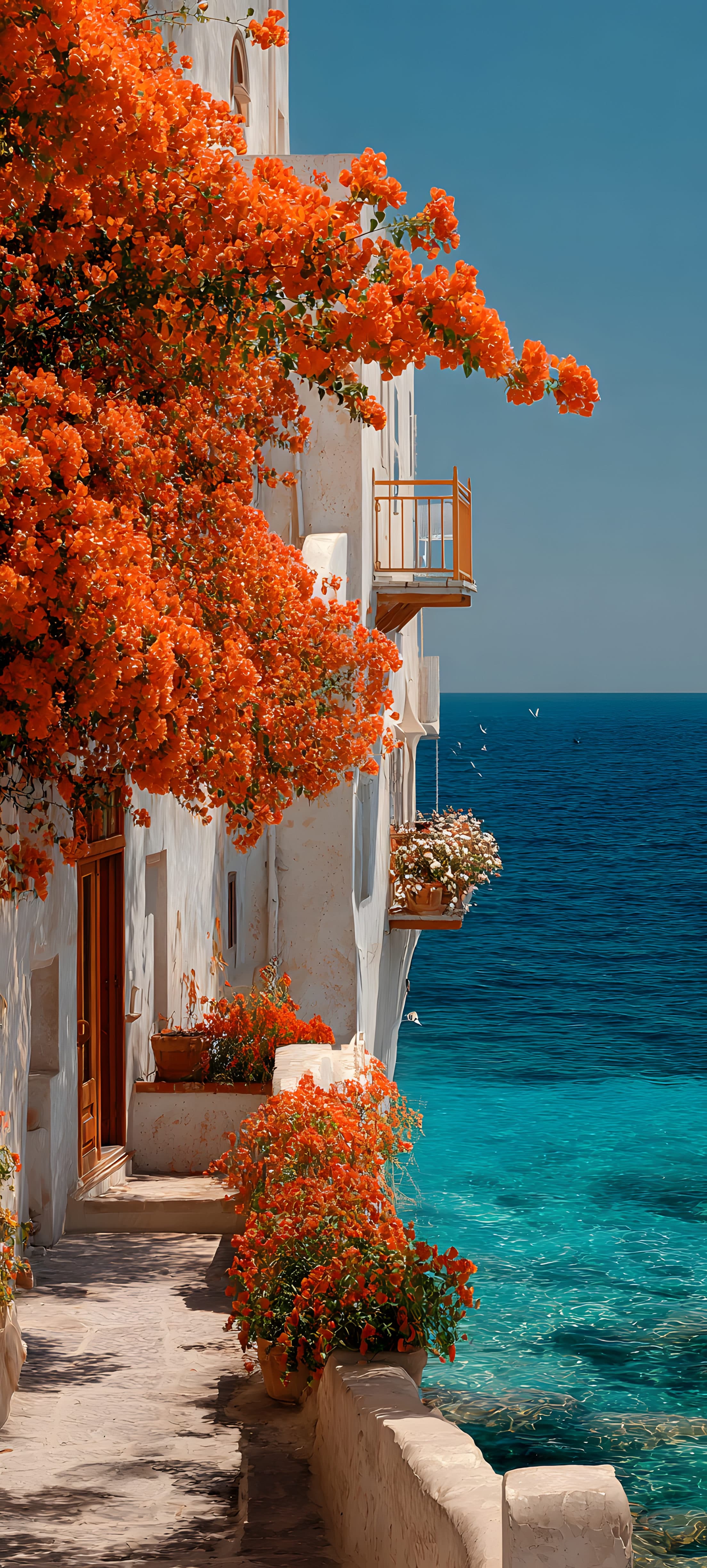 Vibrant orange bougainvillea flowers on a white building overlooking a turquoise sea with a clear blue sky.